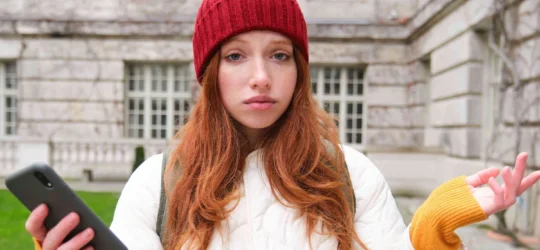 portrait confused redhead girl with smartphone shrugs shoulders with puzzled clueless face 540x250